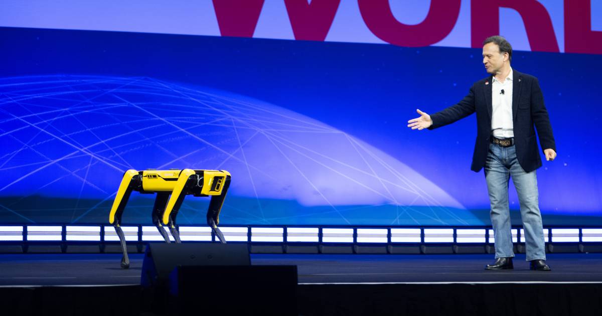 A imagem mostra Gian Paolo Bassi, vice-presidente sênior da Dassault Systèmes, em um palco durante um evento. Ele está vestindo um blazer preto, camisa branca e jeans, e gesticula em direção a um robô quadrúpede amarelo, que parece ser o Spot, desenvolvido pela Boston Dynamics. O fundo apresenta uma grande tela azul com gráficos digitais, sugerindo um ambiente tecnológico