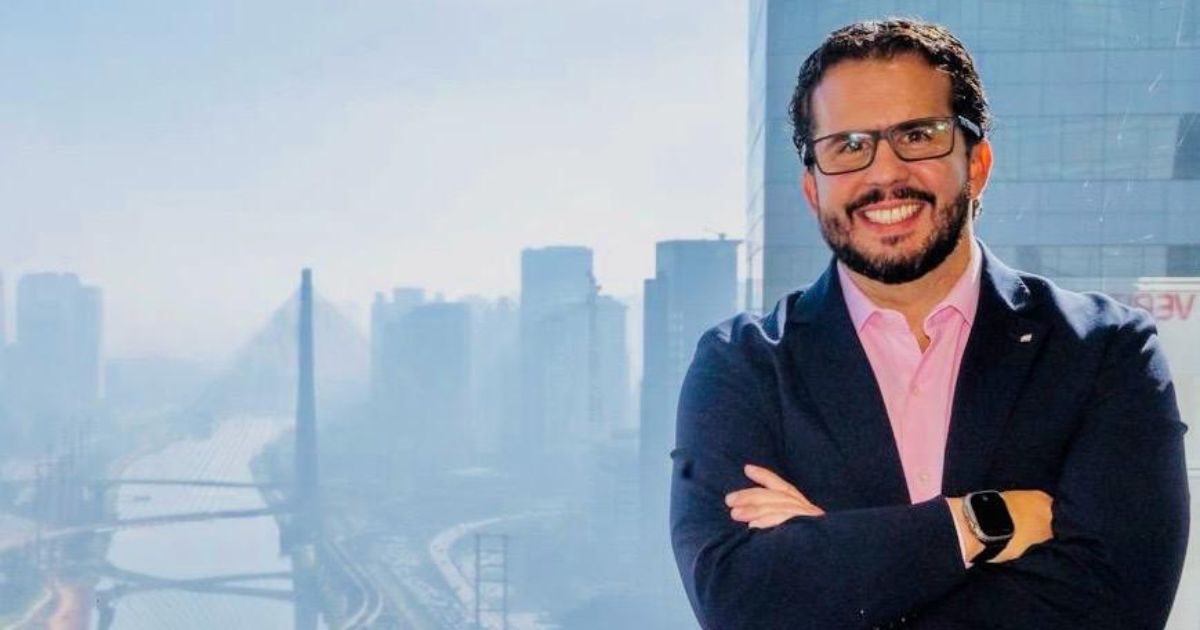 Homem sorridente com óculos e blazer azul posando em frente a uma janela com vista para a cidade de São Paulo, destacando a Ponte Estaiada e prédios ao fundo.