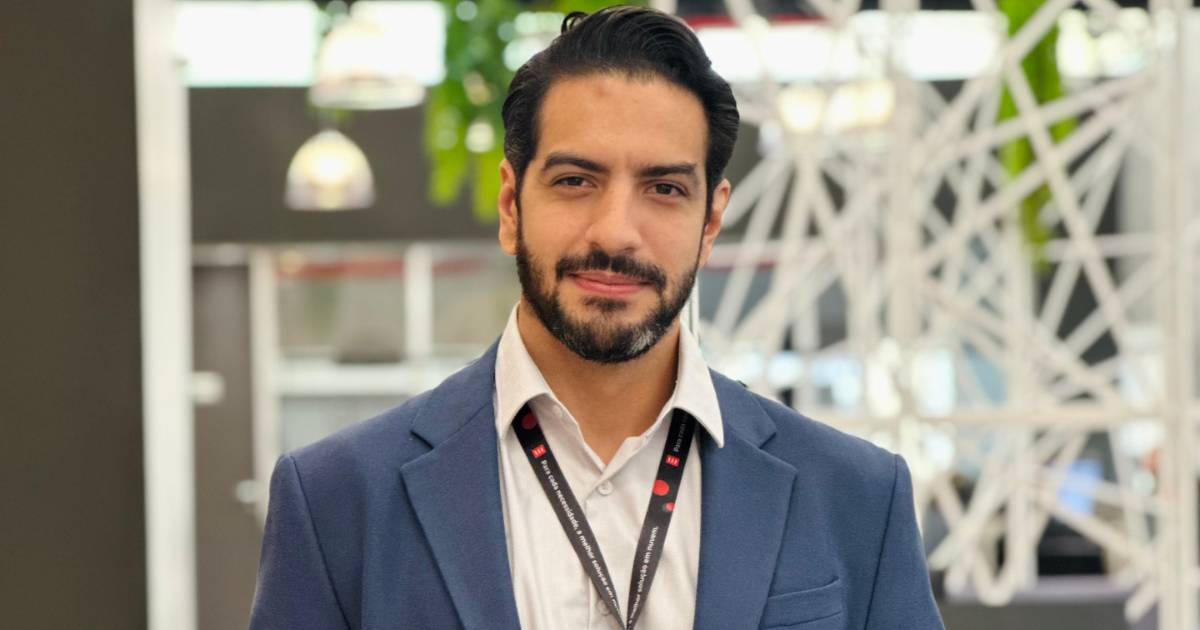 Kleber Rangel, novo diretor de Tecnologia da Eveo, posando para uma foto em um ambiente corporativo moderno. Ele veste um blazer azul sobre uma camisa branca e usa um crachá preto. O fundo desfocado apresenta elementos arquitetônicos e iluminação pendente