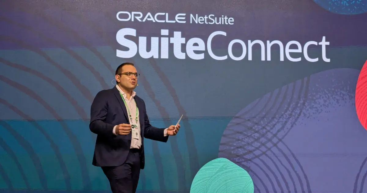 Imagem de Gustavo Moussalli, vice-presidente da NetSuite para a América Latina, durante o evento SuiteConnect da Oracle NetSuite. Ele está no palco, vestindo um terno azul escuro, camisa clara e crachá, segurando um dispositivo enquanto faz sua apresentação. O fundo apresenta um grande painel digital com o nome do evento e elementos gráficos modernos