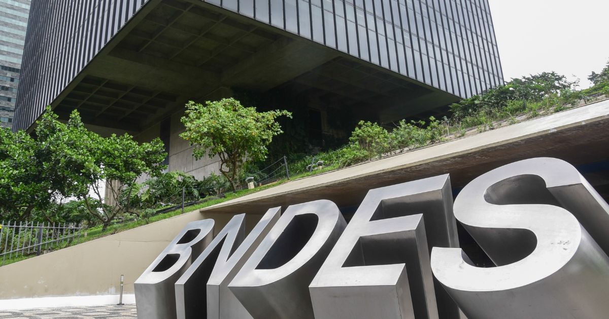 Fotografia da fachada do edifício do BNDES (Banco Nacional de Desenvolvimento Econômico e Social), com um letreiro metálico grande exibindo a sigla "BNDES" em destaque no primeiro plano. O prédio moderno, com arquitetura robusta e linhas retas, está cercado por áreas verdes e árvores. A perspectiva da foto enfatiza a grandiosidade da construção, com um céu nublado ao fundo.