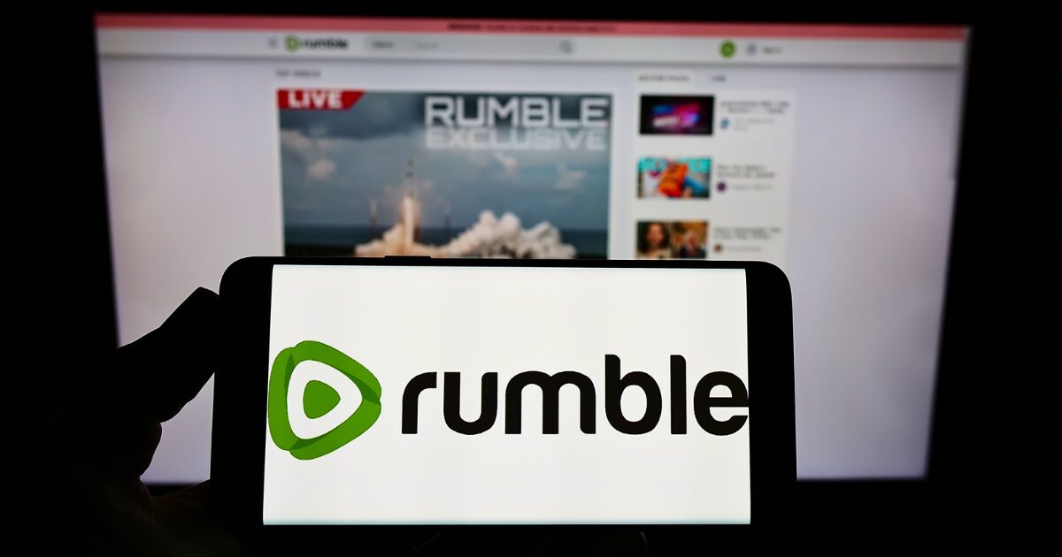 Tela de celular exibindo o logotipo do Rumble em primeiro plano, enquanto, ao fundo, aparece a página inicial da plataforma de vídeos Rumble em um monitor.