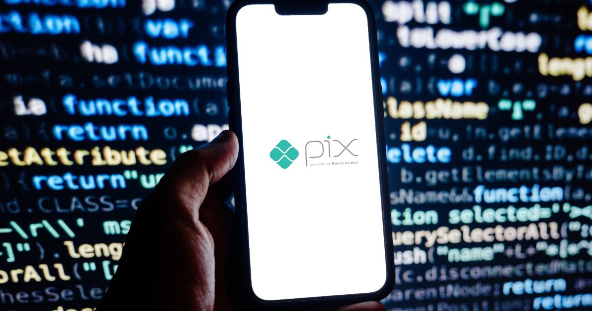 Uma mão segura um smartphone com a tela branca exibindo o logotipo do Pix, sistema de pagamentos do Banco Central do Brasil. O logotipo consiste em um ícone verde ao lado da palavra "Pix" escrita em letras cinza e verdes. Ao fundo, há uma tela com códigos de programação destacados em cores como azul, amarelo e branco, sugerindo um ambiente digital ou de segurança cibernética. A imagem transmite a ideia de tecnologia, segurança e transações financeiras digitais.
