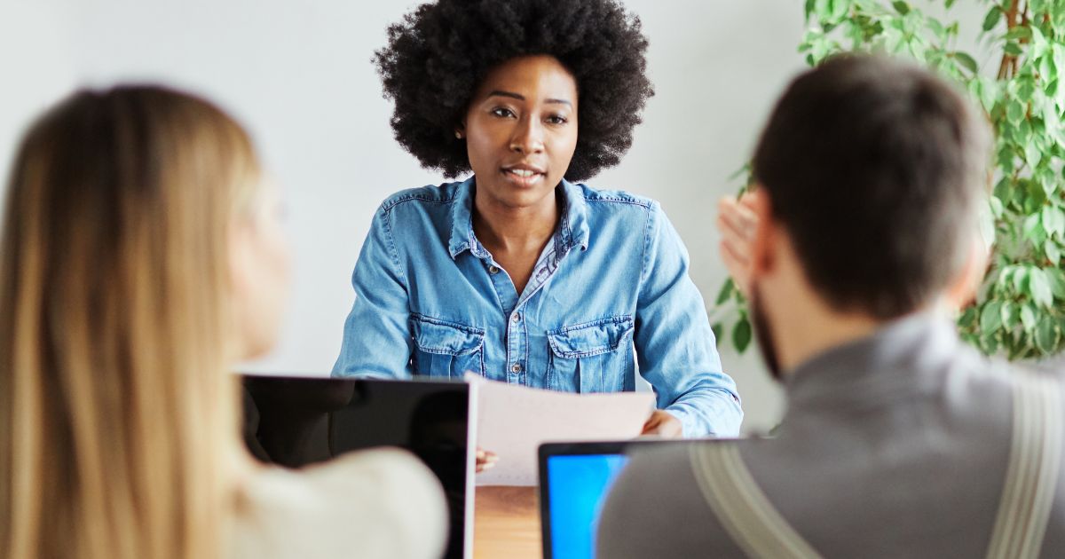 "Uma mulher negra com cabelo afro, vestindo uma camisa jeans, está sentada à frente de duas pessoas em um ambiente de escritório. Ela segura um papel e parece estar participando de uma entrevista ou reunião. As outras duas pessoas, um homem e uma mulher, estão sentadas de costas para a câmera, com laptops abertos à sua frente. O ambiente tem uma decoração moderna, com uma planta verde ao fundo.