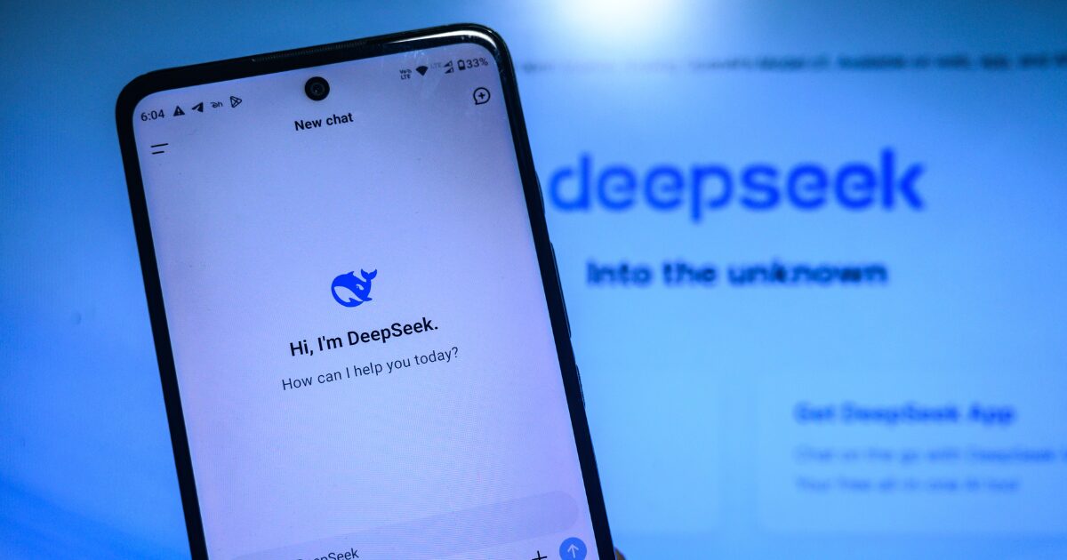 Uma imagem mostrando a tela de um smartphone com o aplicativo da startup "DeepSeek" aberto. Na tela, é exibida a mensagem: "Hi, I'm DeepSeek. How can I help you today?" com o logotipo do aplicativo, que representa um golfinho estilizado em azul. Ao fundo, desfocado, está o logotipo maior do "DeepSeek" junto com o texto "Into the unknown".