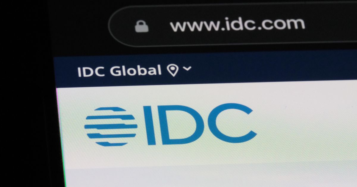 Captura de ecrã de um site mostrando o logótipo da IDC (International Data Corporation). O logótipo consiste em letras azuis 'IDC' ao lado de um ícone circular com linhas horizontais. Acima, há um menu com o texto 'IDC Global' e um ícone de localização. No topo da imagem, a barra de endereço do navegador exibe 'www.idc.com' com um cadeado indicando conexão segura (crescimento)