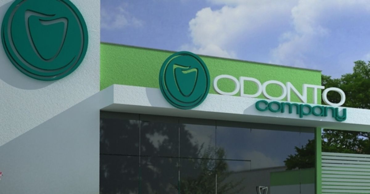 A imagem mostra a fachada de uma clínica odontológica da rede "Odonto Company". A construção tem um design moderno, com tons predominantes de verde e branco. O logotipo da empresa, que apresenta um dente estilizado dentro de um círculo, está em destaque na parede e ao lado do nome "Odonto Company" em letras grandes. A entrada possui uma grande área envidraçada, refletindo árvores e o céu azul ao fundo