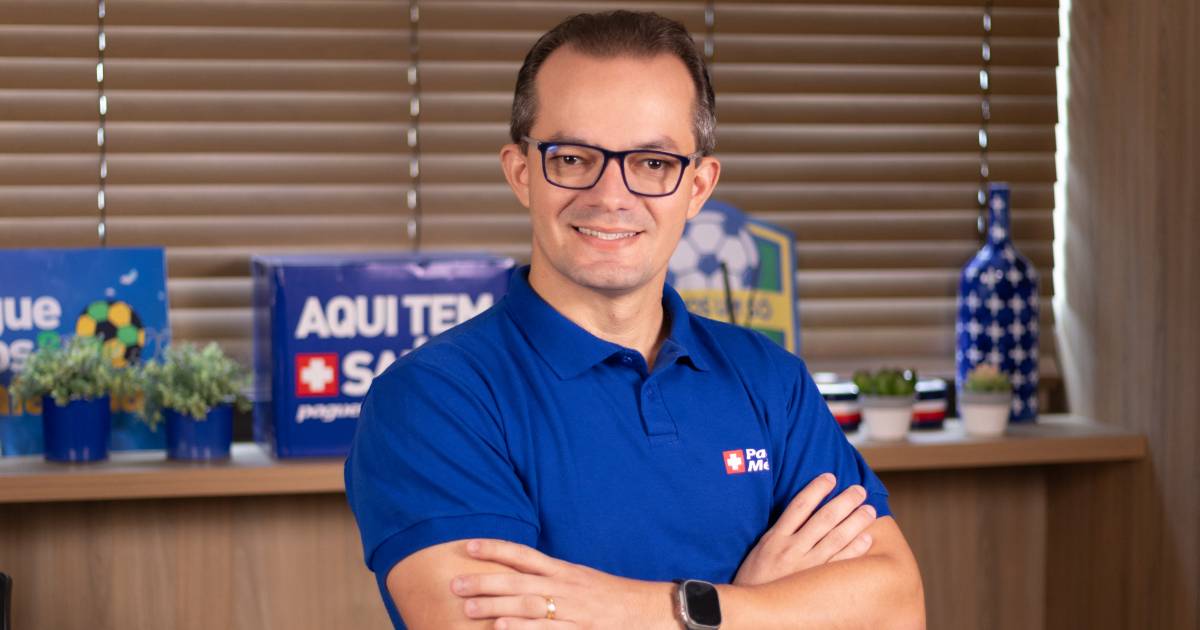 Plínio Girão, diretor de Dados da Pague Menos, sorrindo e posando com os braços cruzados. Ele veste uma camisa azul com o logotipo da empresa, com um ambiente corporativo ao fundo