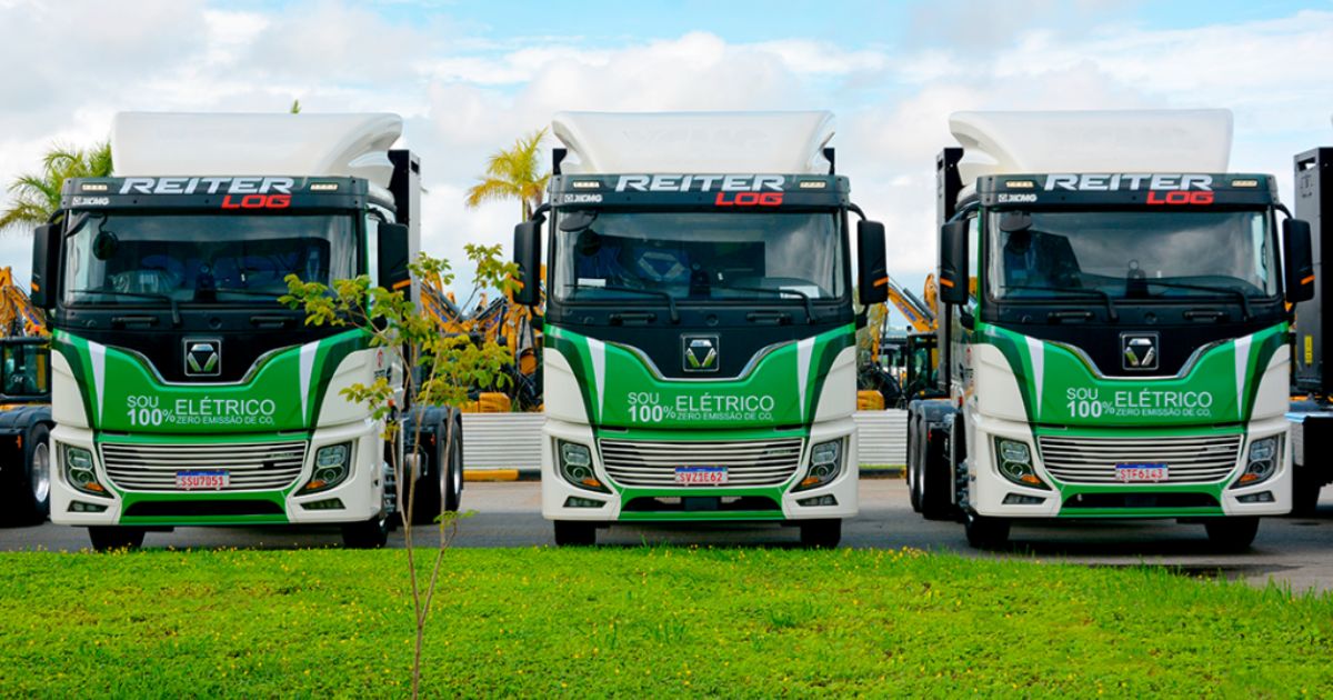 Três caminhões elétricos da Reiter Log alinhados lado a lado, com design verde e branco e a mensagem 'Sou 100% elétrico, zero emissão de CO₂' estampada na frente. O cenário ao fundo inclui palmeiras, máquinas pesadas e um céu parcialmente nublado (caminhão elétrico, suzano, reiter log, xcmg)