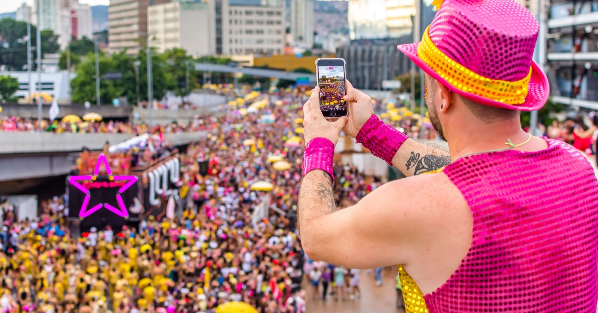 A imagem mostra um homem vestido com roupas chamativas em tons de rosa e amarelo, incluindo um chapéu brilhante e acessórios combinando, segurando um smartphone e registrando a cena à sua frente. Ele está em uma posição elevada, filmando ou tirando fotos de um grande bloco de carnaval com uma multidão vestida majoritariamente de amarelo, ocupando uma avenida urbana. O cenário tem prédios altos ao fundo, um trio elétrico decorado com uma estrela neon rosa e diversas pessoas segurando guarda-sóis amarelos. A atmosfera vibrante sugere um evento festivo de carnaval em uma grande cidade (golpes)