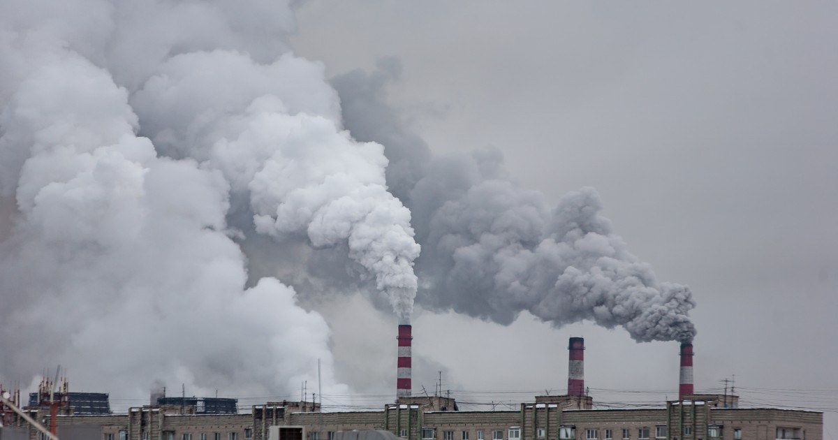 Imagem de chaminés de uma usina industrial emitindo grandes quantidades de fumaça branca e cinza em direção ao céu nublado. Abaixo das chaminés, é possível ver prédios residenciais ou industriais. A cena destaca a poluição atmosférica e o impacto ambiental causado pelas emissões (organizações)