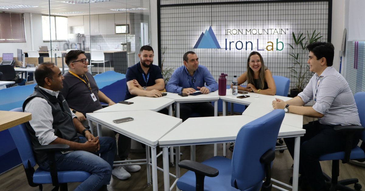 A imagem mostra Sabrina Melo, diretora da Iron Mountain Brasil, reunida com sua equipe no espaço de inovação da empresa, o Iron Lab. O ambiente é moderno e colaborativo, com móveis modulares e tons de azul. A equipe está sentada ao redor de mesas dispostas em formato circular, demonstrando um clima descontraído e interativo. Ao fundo, há um letreiro iluminado com o nome do Iron Lab, reforçando o caráter inovador do espaço.