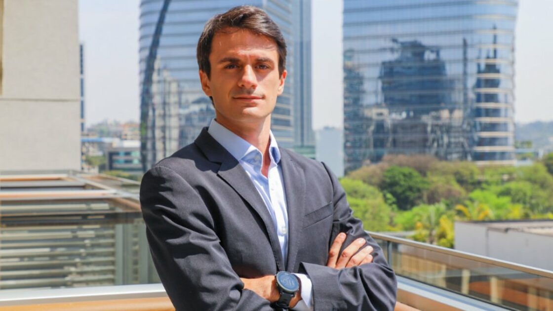 A imagem mostra Juranilson Santos, diretor de logística da WDC, posando para uma foto profissional em uma varanda com vista para prédios modernos e áreas verdes ao fundo. Ele está vestindo um terno escuro com camisa azul clara, tem os braços cruzados e exibe uma expressão séria e confiante. A iluminação natural destaca seu rosto e cria um contraste equilibrado com o fundo urbano. (Itajaí)