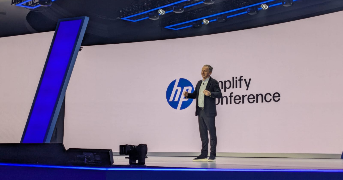 A imagem mostra Enrique Lores, CEO da HP Inc., no palco da HP Amplify Conference. Ele está vestido com um terno escuro e uma camisa clara, gesticulando enquanto discursa para a plateia. O palco tem um design moderno, com iluminação azul e um grande telão ao fundo exibindo o logotipo da HP e o nome do evento