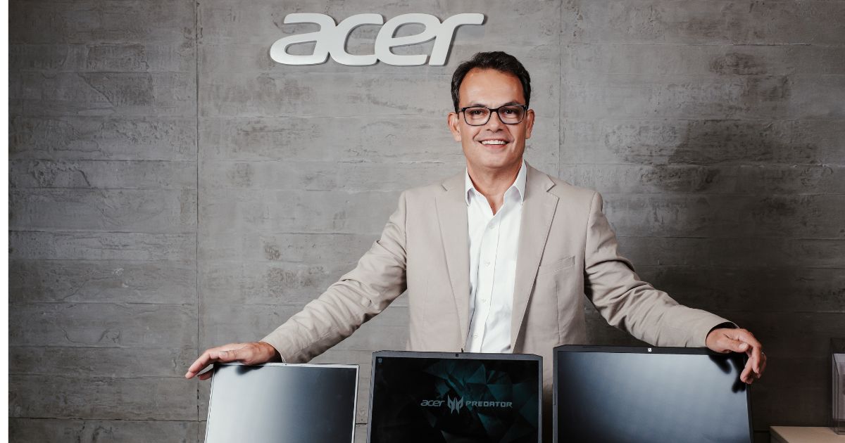 Germano Couy, gerente-geral da Acer Brasil e América Latina, posando sorridente em um ambiente corporativo da Acer. Ele veste um terno bege e uma camisa branca, apoiando as mãos sobre três notebooks da marca, incluindo um modelo da linha Predator. O logotipo da Acer está visível na parede de concreto ao fundo.