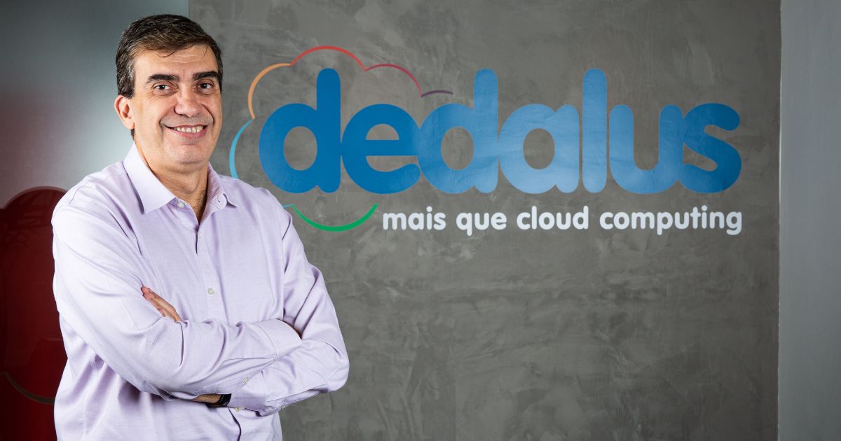 Maurício Fernandes, CEO da Dedalus, posa sorridente com os braços cruzados em frente ao logo da empresa. O fundo é uma parede cinza com o nome 'dedalus' em letras azuis e a frase 'mais que cloud computing' em branco. Foto: Divulgação/Dedalus