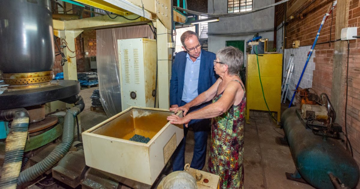 O presidente da ABDI, Ricardo Cappelli, visita a fábrica da ABL Plásticos e conversa com Dulce Brenner. Ele veste um terno azul e observa atentamente enquanto Dulce, trajando um vestido estampado, explica sobre o maquinário. O ambiente industrial apresenta equipamentos grandes, tubulações e paredes de tijolos aparentes, demonstrando um espaço de produção. Em primeiro plano, uma máquina contém pequenos grânulos plásticos, indicando o processo produtivo da fábrica (ABDI, Recupera Indústria RS, Senai-RS, Fiergs, enchentes)