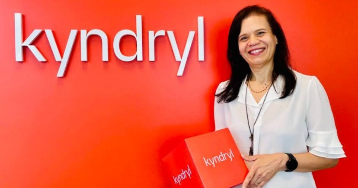 Uma mulher de cabelos escuros e longos, sorridente, veste uma blusa branca e segura uma caixa vermelha com a marca 'Kyndryl' impressa. Atrás dela, há uma parede vermelha com o nome 'Kyndryl' em letras brancas tridimensionais. O fundo vibrante contrasta com a roupa clara da mulher, destacando a identidade visual da marca.