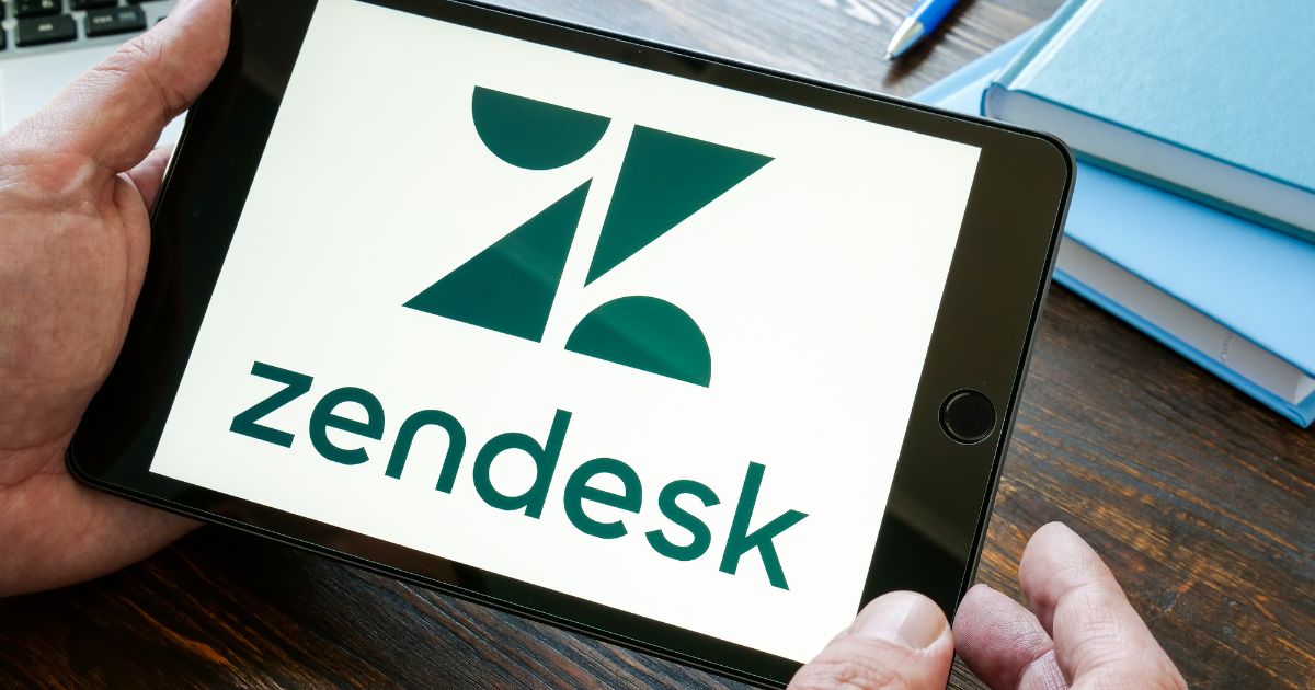 Um tablet sendo segurado por uma pessoa, exibindo o logotipo do Zendesk em uma tela branca. O logotipo é verde e estilizado, com o nome "Zendesk" abaixo. No fundo, há uma mesa de madeira, alguns cadernos e uma caneta azul.