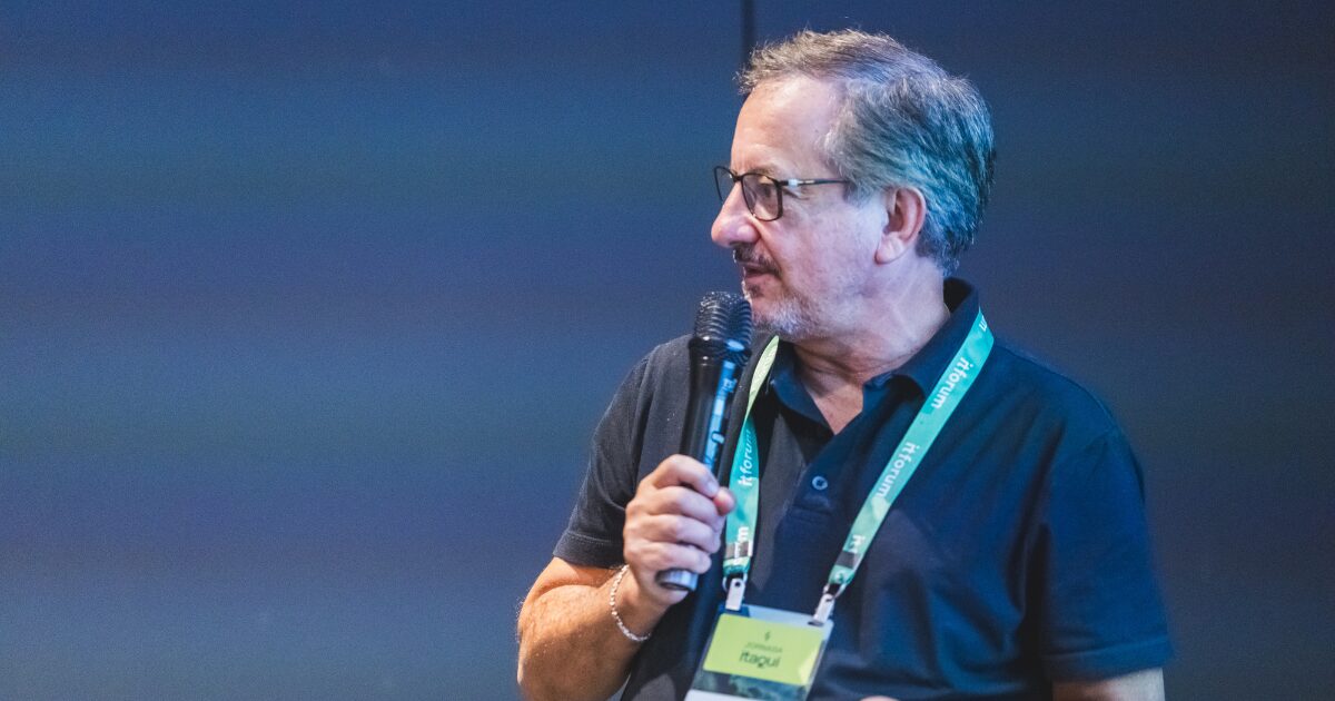 Homem de meia-idade, com barba grisalha e óculos, segura um microfone enquanto fala durante um evento. Ele usa uma camisa polo preta e um crachá verde suspenso no pescoço com a inscrição "Itaqui" e "IT Forum". O fundo da imagem é um painel azul desfocado. Nelson Campelo, CEO da Atos para a América Latina durante o Jornada Itaqui