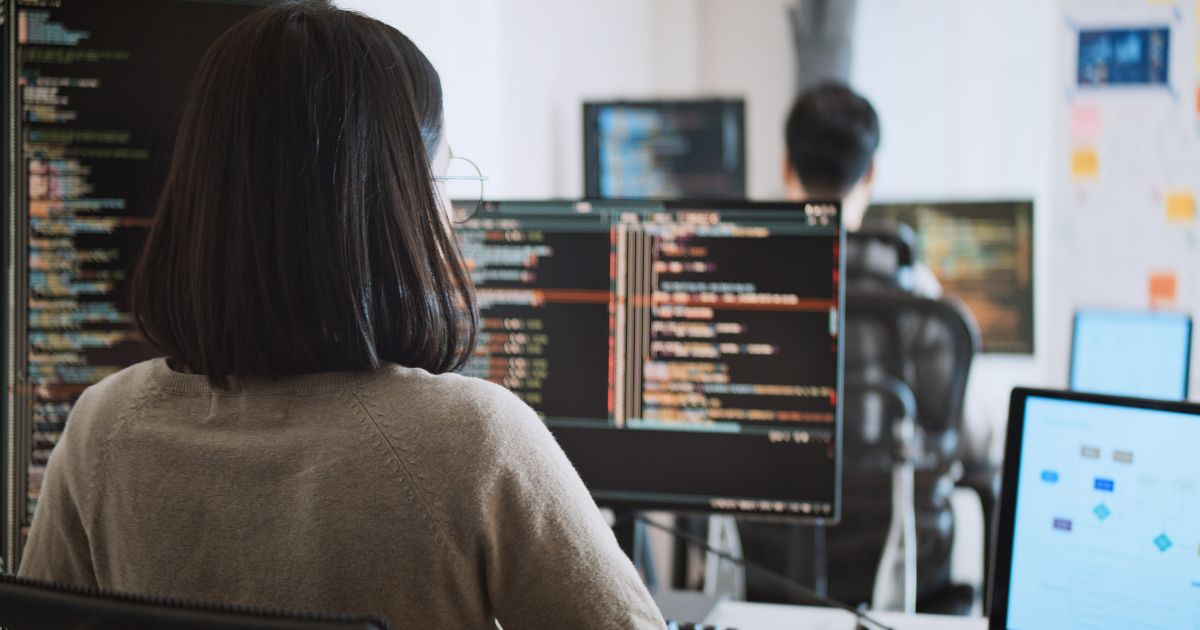 Uma mulher de cabelo escuro e óculos está sentada em frente a vários monitores de computador, trabalhando com código de programação. A tela principal exibe linhas de código coloridas em um ambiente de desenvolvimento. No fundo, outro programador, um homem de cabelo curto, também trabalha em seu computador, e há um quadro branco com anotações e diagramas. O ambiente parece ser um escritório moderno de tecnologia ou uma startup.