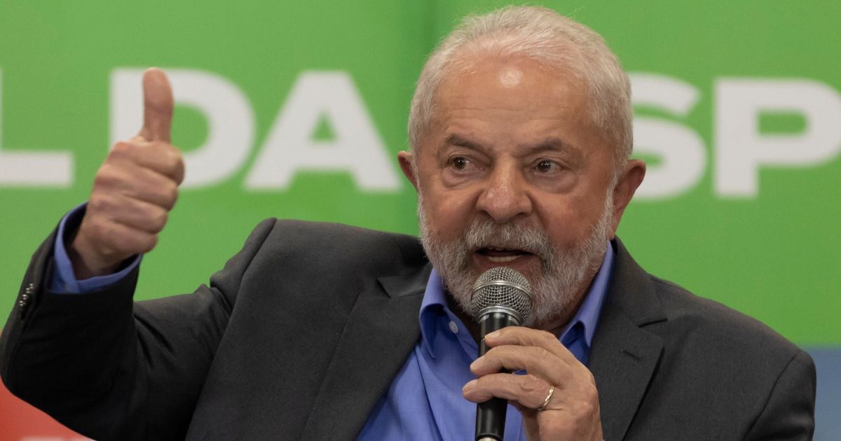 Um homem idoso com barba grisalha segura um microfone enquanto fala. Ele veste um blazer escuro sobre uma camisa azul e faz um gesto de positivo com a mão esquerda. O fundo é verde com letras brancas parcialmente visíveis. Ele tem uma expressão determinada e está olhando para alguém ou algo fora do quadro.