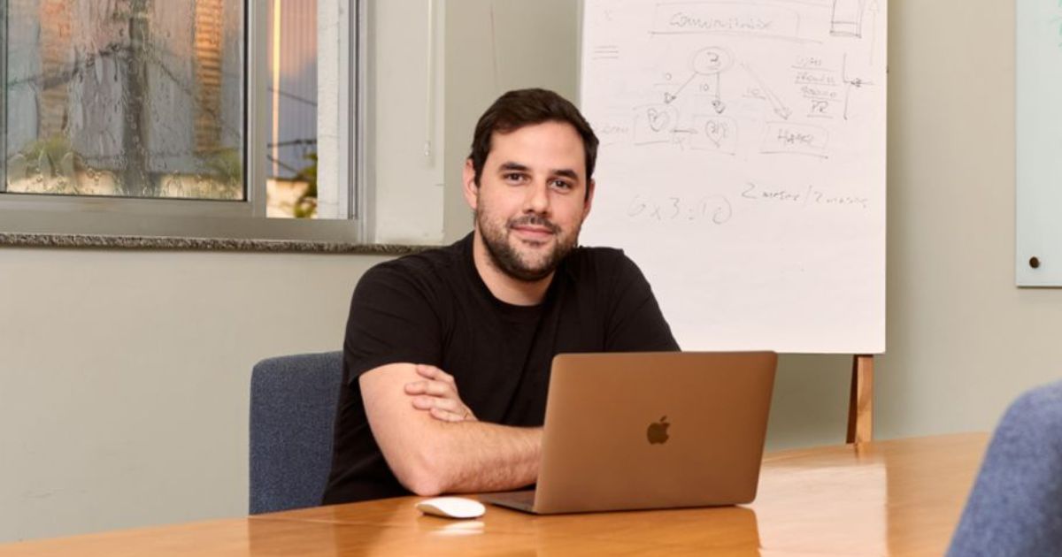 Rodrigo Stoqui, country manager da Tldv, sentado à mesa de uma sala de reunião. Ele veste uma camisa preta e tem um laptop da Apple à sua frente. Atrás dele, há um quadro branco com anotações e diagramas. A sala tem uma janela ao fundo, com vidros levemente embaçados