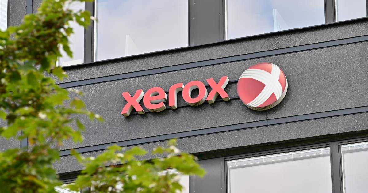 A imagem mostra a fachada de um edifício com o logotipo da empresa Xerox fixado na parede externa. O logotipo é composto pelo nome "Xerox" em letras vermelhas e um símbolo circular vermelho com linhas brancas cruzadas. A construção tem um design moderno, com painéis escuros e janelas de vidro refletindo o céu. Na parte frontal da imagem, há folhas verdes de uma árvore parcialmente desfocadas, adicionando um toque natural à cena.