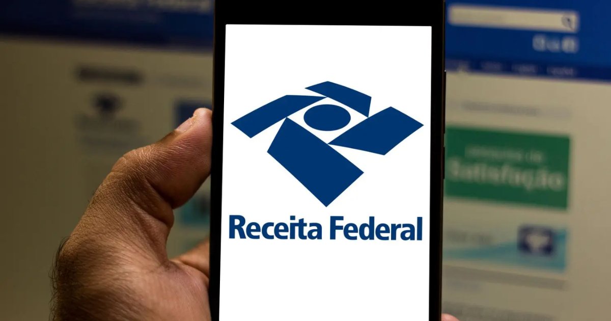 A imagem mostra uma mão segurando um smartphone com a tela exibindo o logotipo da Receita Federal do Brasil, que consiste em um símbolo geométrico azul e o nome da instituição escrito abaixo. No fundo, desfocado, há um site acessado em um computador, possivelmente relacionado à Receita Federal, com partes do layout visíveis, incluindo uma barra de pesquisa no topo e um botão verde com texto parcialmente legível (imposto de renda, irpf, serpro, receita federal)