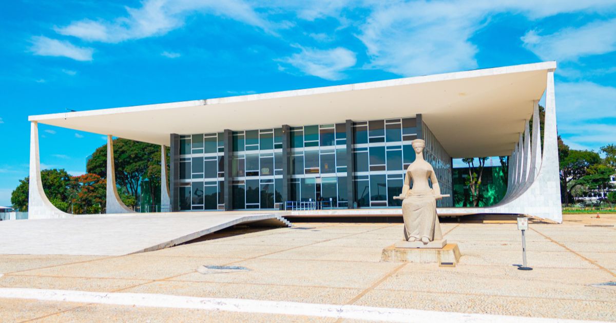 **Descrição alternativa (alt text):** Edifício moderno de arquitetura imponente, com fachada de vidro e linhas retas, coberto por uma grande laje branca sustentada por colunas curvas nas extremidades. Em frente ao prédio, há uma ampla praça de concreto claro, quase sem pessoas, e uma escultura de pedra representando uma figura sentada sobre um pedestal. O céu está azul com algumas nuvens brancas, e há árvores verdes ao redor, especialmente na lateral esquerda e ao fundo do edifício. A iluminação é forte e natural, sugerindo um dia ensolarado. (STF)