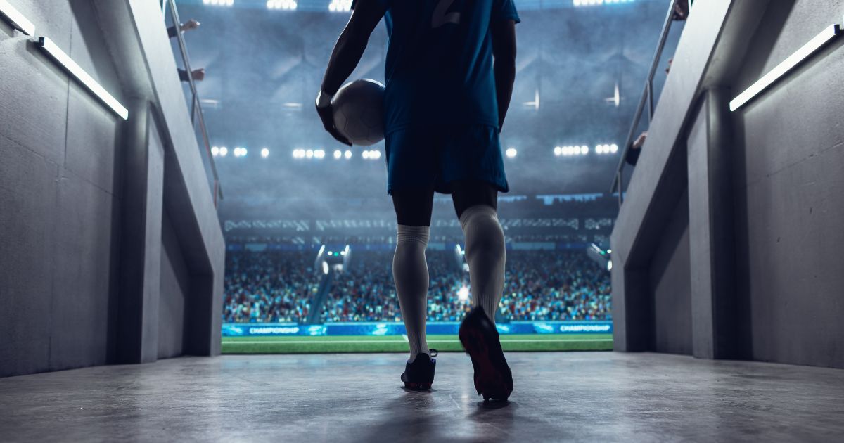 Sport adota análise de dados para gestão de atletas e contratações 4 **Descrição alternativa (alt text):** Vista a partir de um túnel de acesso ao campo, mostrando uma pessoa vestindo uniforme esportivo azul caminhando em direção a um estádio iluminado, segurando uma bola de futebol sob o braço. A cena é capturada por trás, com foco nas pernas e nas chuteiras. O túnel possui paredes laterais com corrimãos e iluminação artificial. Ao fundo, um grande estádio aparece cheio de público, com luzes fortes e o campo verde visível na saída do túnel, criando um contraste entre a área interna sombreada e o ambiente externo luminoso. (Sport)